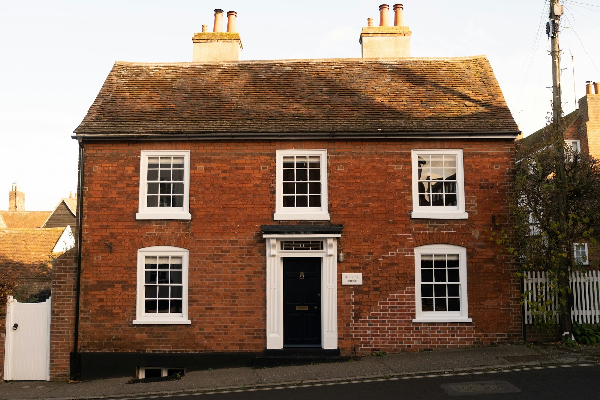 A red-brick house with white sash windows on a British street, in low afternoon sun